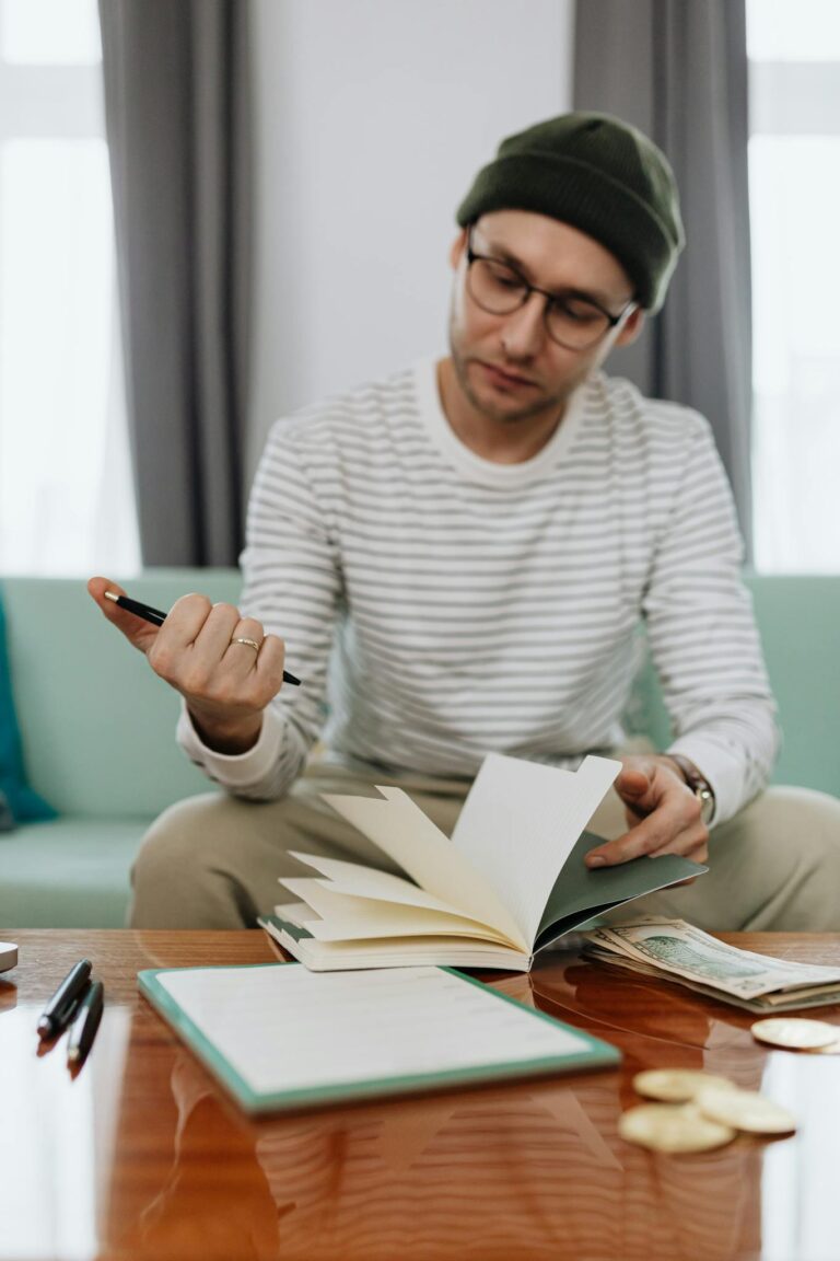 A young man wearing a beanie and glasses reviews his finances sitting on a couch.