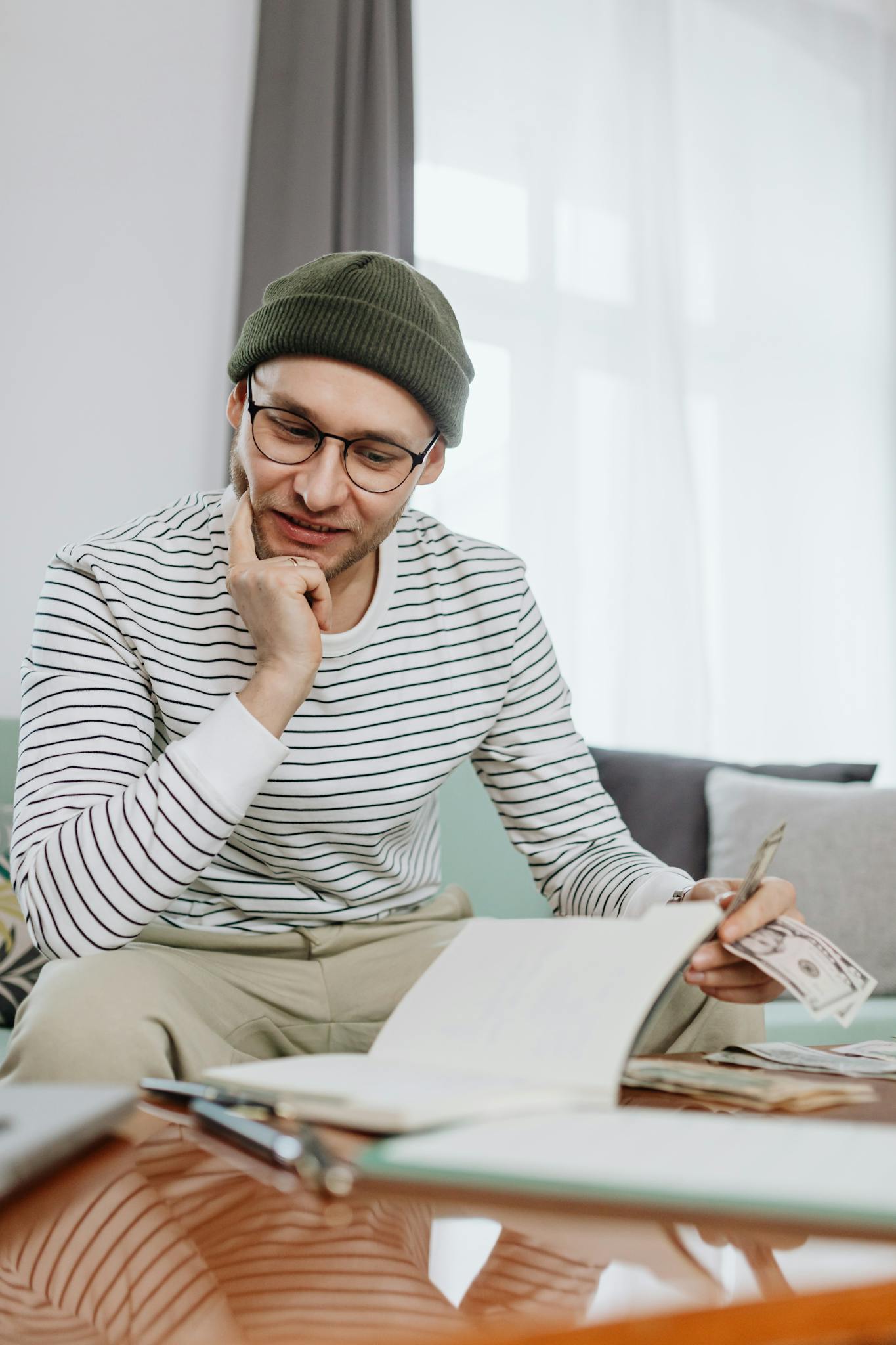 Young man in beanie and glasses reviewing finances with dollar bills and notebook on table.